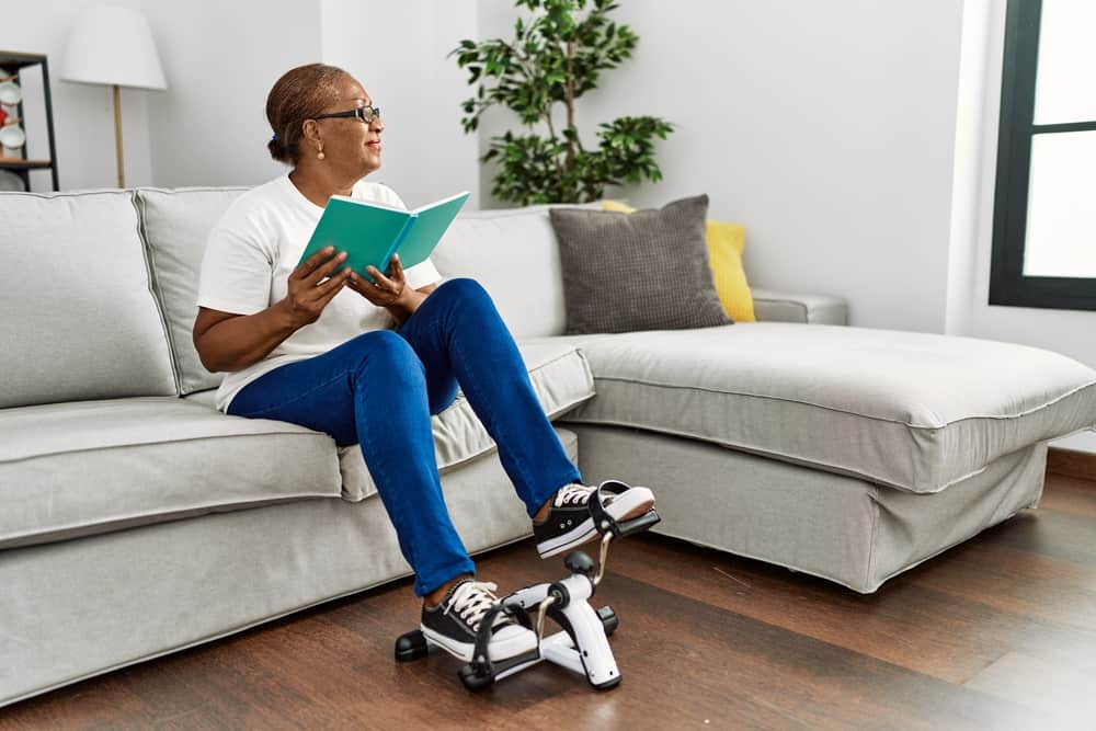 An elderly woman using a mini exercise bike while reading a book in her living room