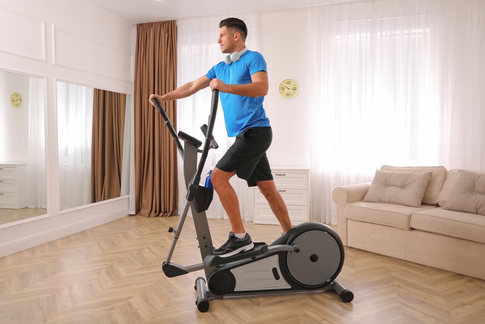 Young man wearing blue top with headphones around his neck working out on his elliptical cross trainer in his home